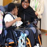 A smiling Support Worker sits beside a child in a wheelchair during the Special People Open Day.